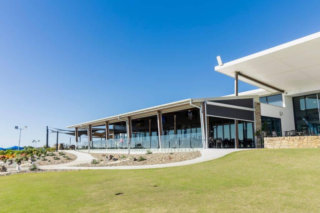 A café with glass walls and black outdoor blinds, set in a landscaped area with a clear blue sky, offering sun protection.