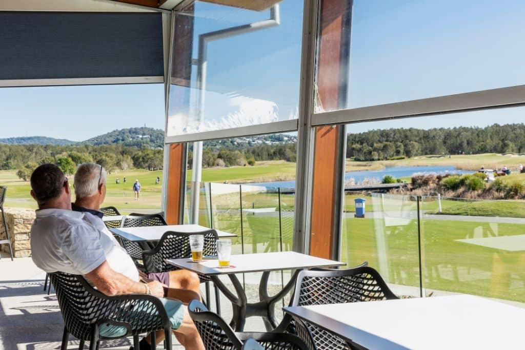 Two men seated in a shaded outdoor area with retractable blinds, enjoying drinks and panoramic views of a scenic golf course.